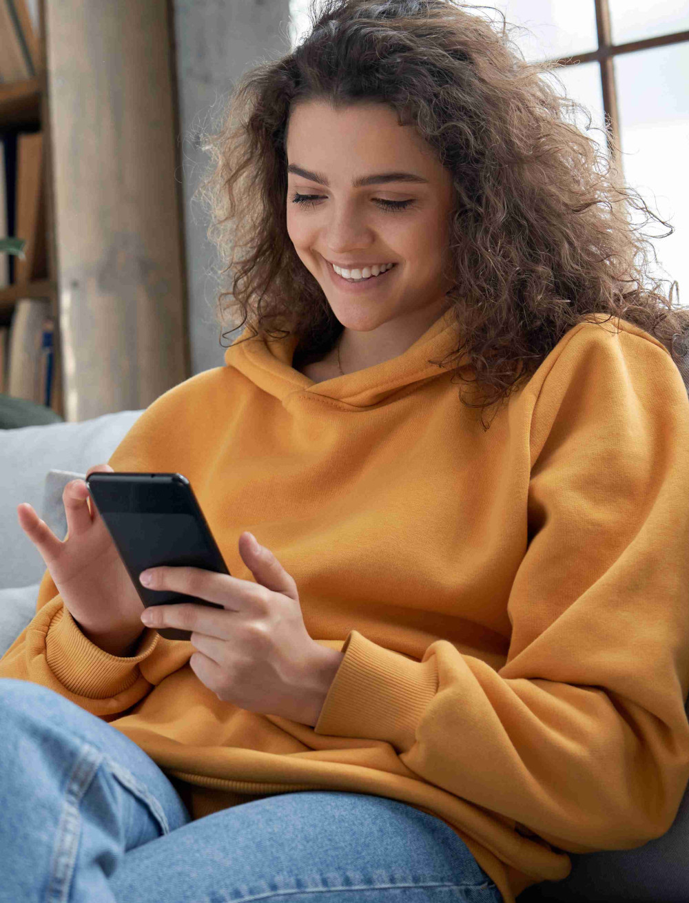 Eine lächelnde junge Frau sitzt gemütlich auf der Couch und nutzt ihr Smartphone.