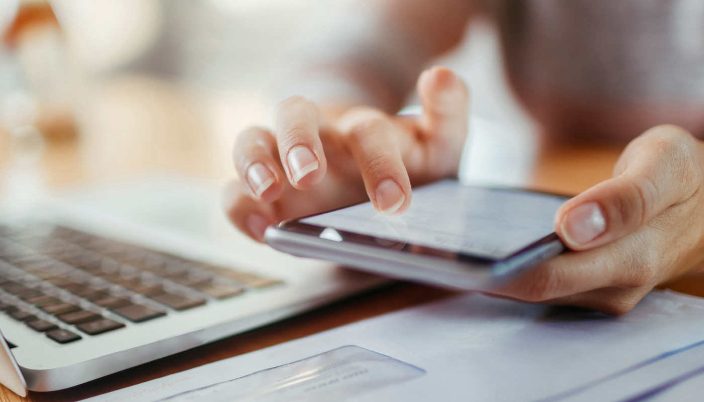 A desk with a laptop and other work materials on it; above it are two hands operating a smartphone