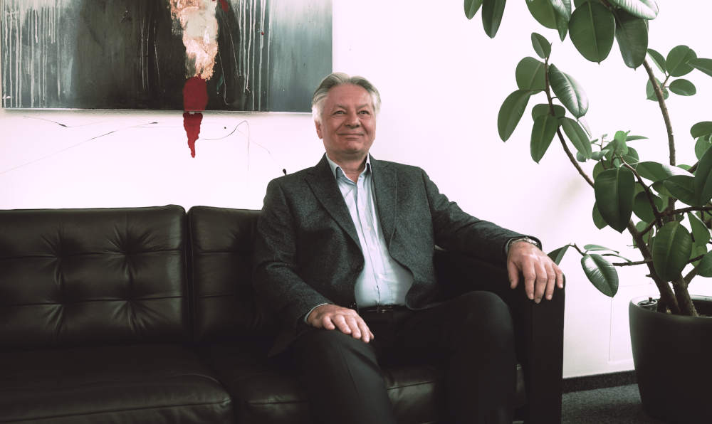 Ein älterer Mann in Businesskleidung sitzt entspannt auf einem schwarzen Ledersofa in einem hellen Büro. Hinter ihm hängt ein abstraktes Gemälde, und rechts steht eine große grüne Zimmerpflanze