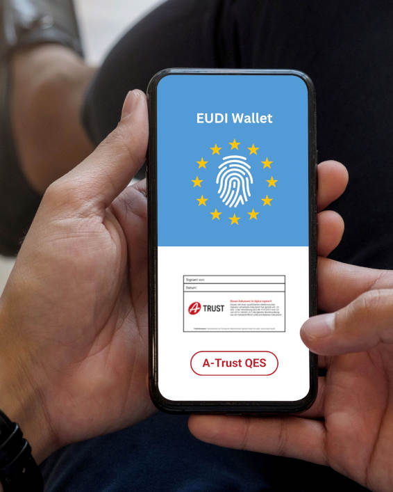 Eine Person hält ein Smartphone; Auf dem oberen Bildschirmbereich steht „EUDI Wallet“ neben einem Fingerabdruck-Symbol in den Farben der Europaflagge; Im unteren Bereich ist die A-Trust Signaturmarke zu sehen sowie ein Button mit der Beschriftung „A-Trust QES“.