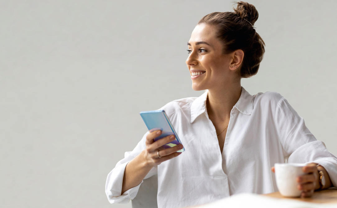 A young, smiling woman sits at a table comfortably; she is using a cell phone while drinking a cup of coffee.