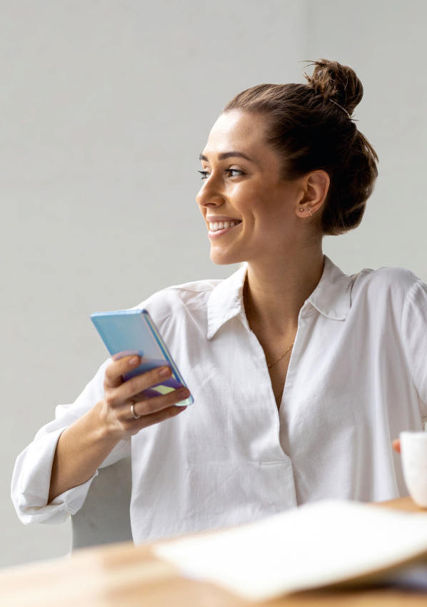 A young, smiling woman sits at a table comfortably; she is using a cell phone while drinking a cup of coffee.