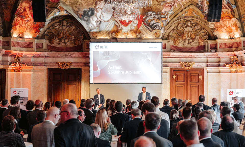 Menschenmenge bei einer feierlichen Veranstaltung in einem prunkvollen Saal mit barocker Decke und Kronleuchter. Zwei Männer im Anzug sprechen auf einer Bühne vor einer Leinwand mit der Aufschrift „A-Trust 25 Jahre Jubiläum“. Im Vordergrund stehen zahlreiche Gäste, einige mit Getränken in der Hand.