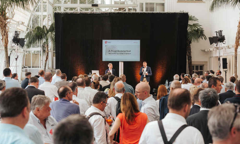 The image shows an event taking place in a large, light-filled room with a glass roof and palm trees in the background. Two men are standing on a stage in front of a black curtain, speaking to a large group of people who are attentively listening. On a screen behind the speakers, the text 'A-Trust Sommerfest - Networking unter Palmen' is displayed. The attendees are mostly dressed in business attire, and the atmosphere appears professional and business-like.