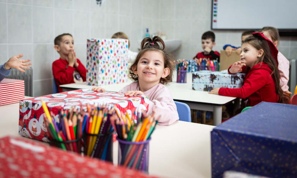Classroom with several children who have received Christmas gift packages; In the foreground, a young girl smiling broadly while holding her present.