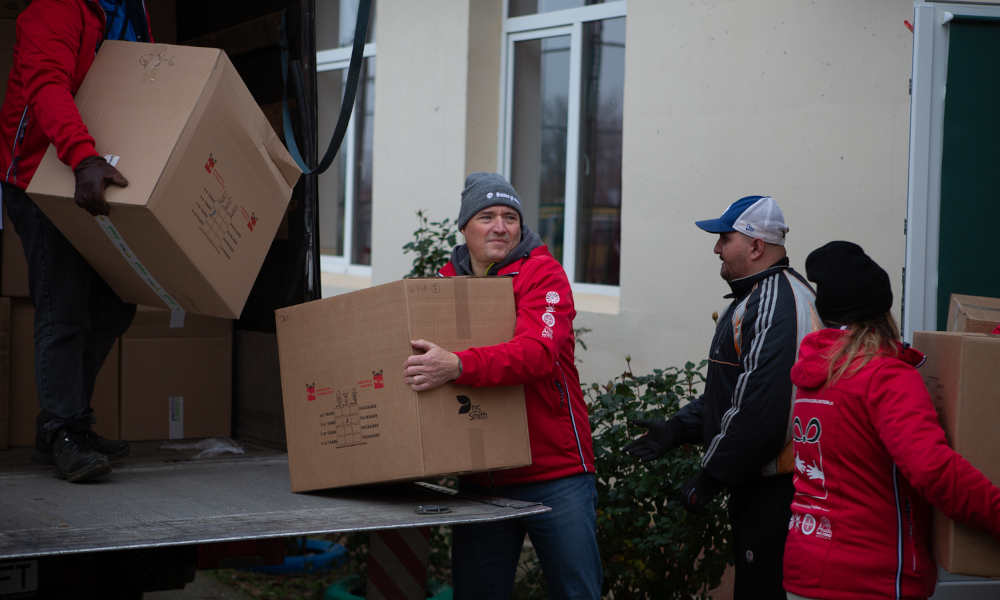 A-Trust Managing Director Sven Glöckner lifting a box of Christmas gift packages from a truck, surrounded by people passing additional boxes.