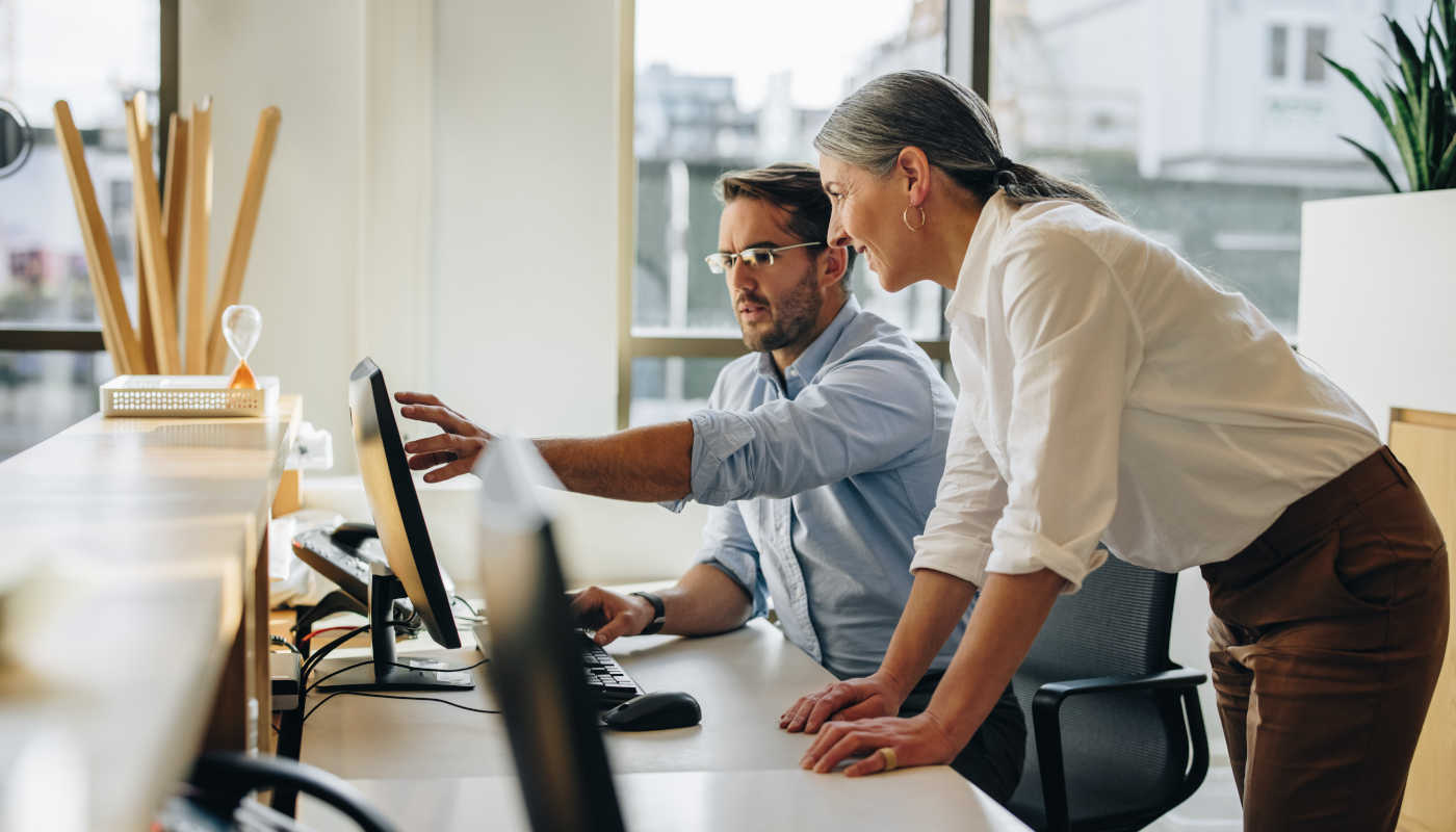 A man shows his colleague something on his screen; she watches intently and smiles.