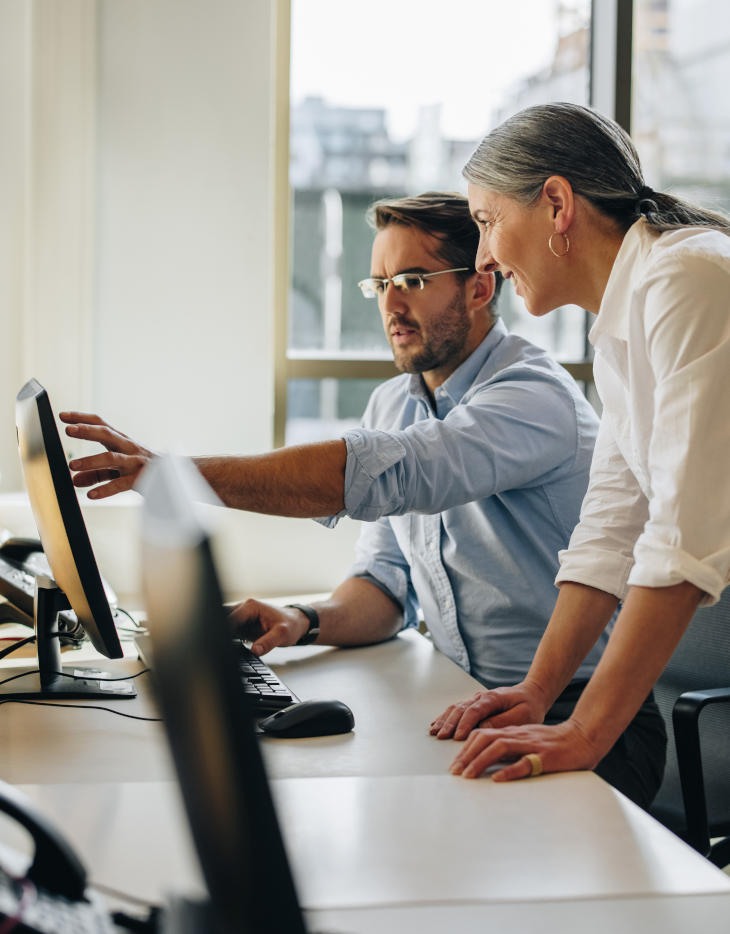 A man shows his colleague something on his screen; she watches intently and smiles.